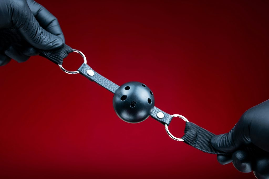 Bâillon BDSM : accessoire pour pimenter ses soirées en couple hands in black gloves holding a round gag in the mouth for erotic bdsm games