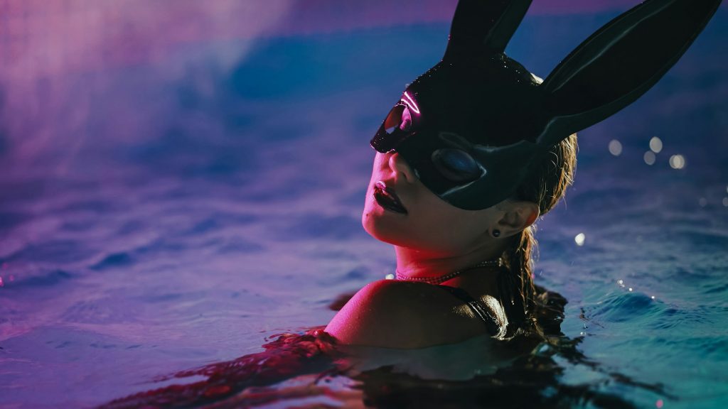 Barre d’écartement : un accessoire de contrainte pour le bondage Sensual woman in leather BDSM mask moving seductively in swimming pool water under neon color light